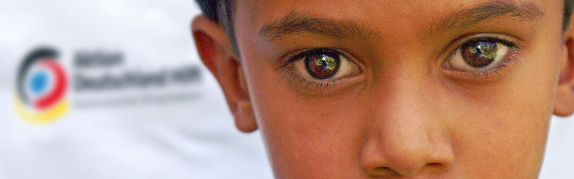 Glossar Humanitäre Hilfe - ein Junge vor dem Logo von Aktion Deutschland Hilft aktion deutschland hilft - bündnis deutscher hilfsorganisationen