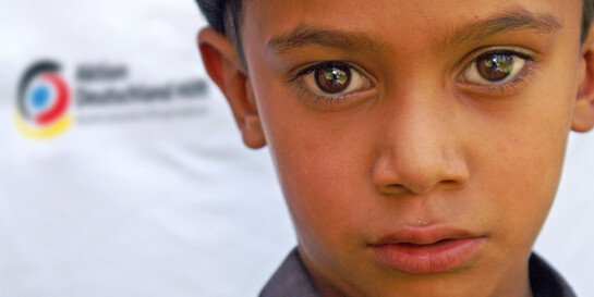 Glossar Humanitäre Hilfe - ein Junge vor dem Logo von Aktion Deutschland Hilft aktion deutschland hilft - bündnis deutscher hilfsorganisationen